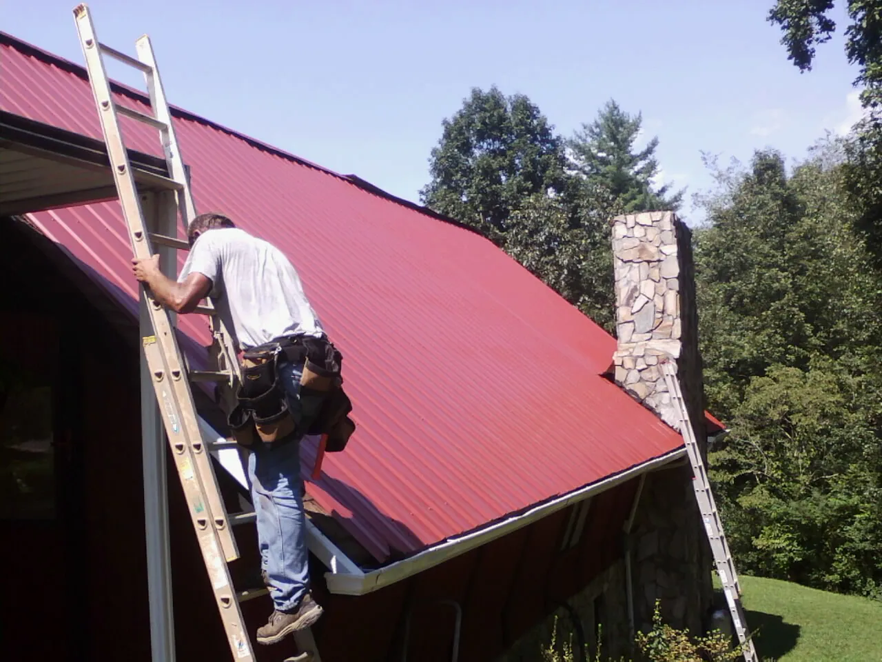 Roofer on a ladder beside a red metal roof and stone chimney — Soffit & Fascia Repair by Roofing Brothers in Alameda